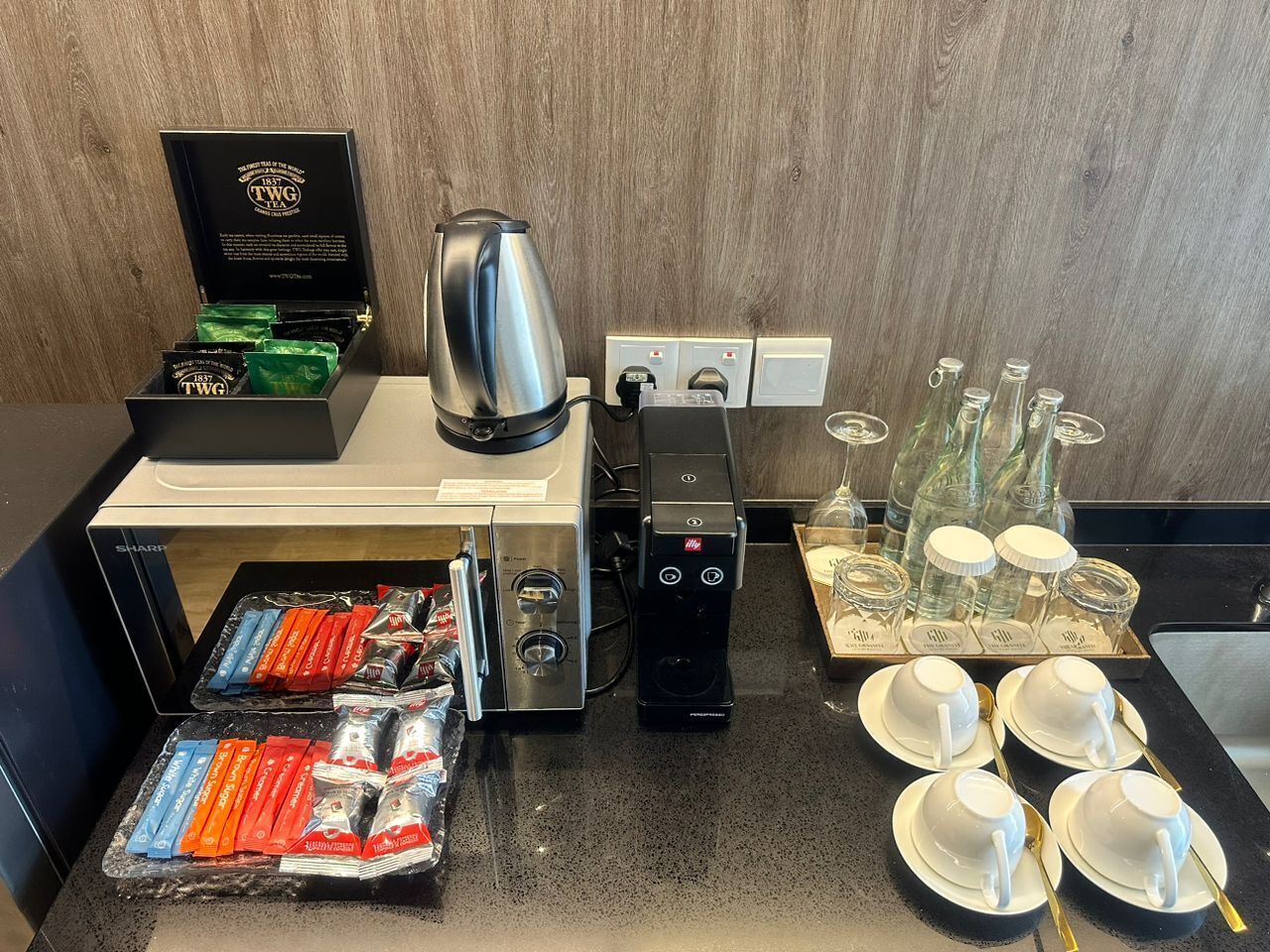 A coffee station with a coffee maker, a box of tea, and a tray of cups and saucers.