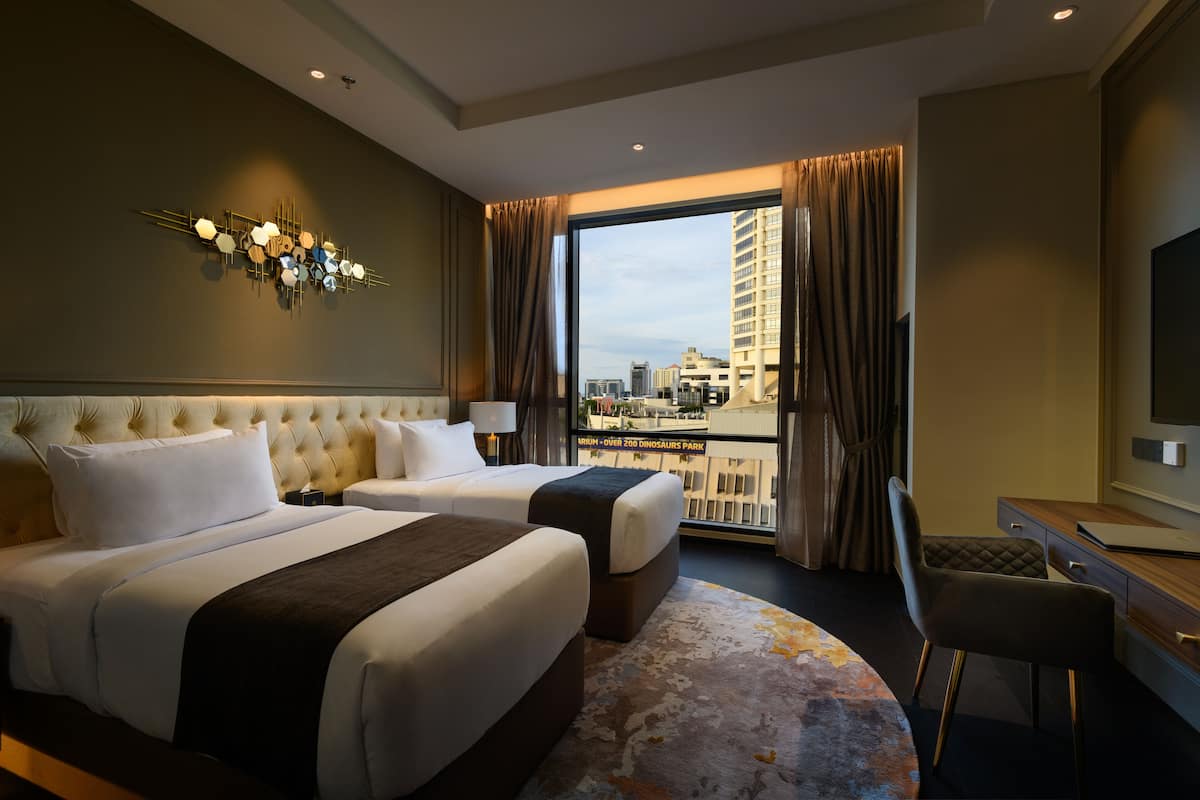 A hotel room with two beds, a desk, a chair, a large window with curtains, and a wall-mounted light fixture.