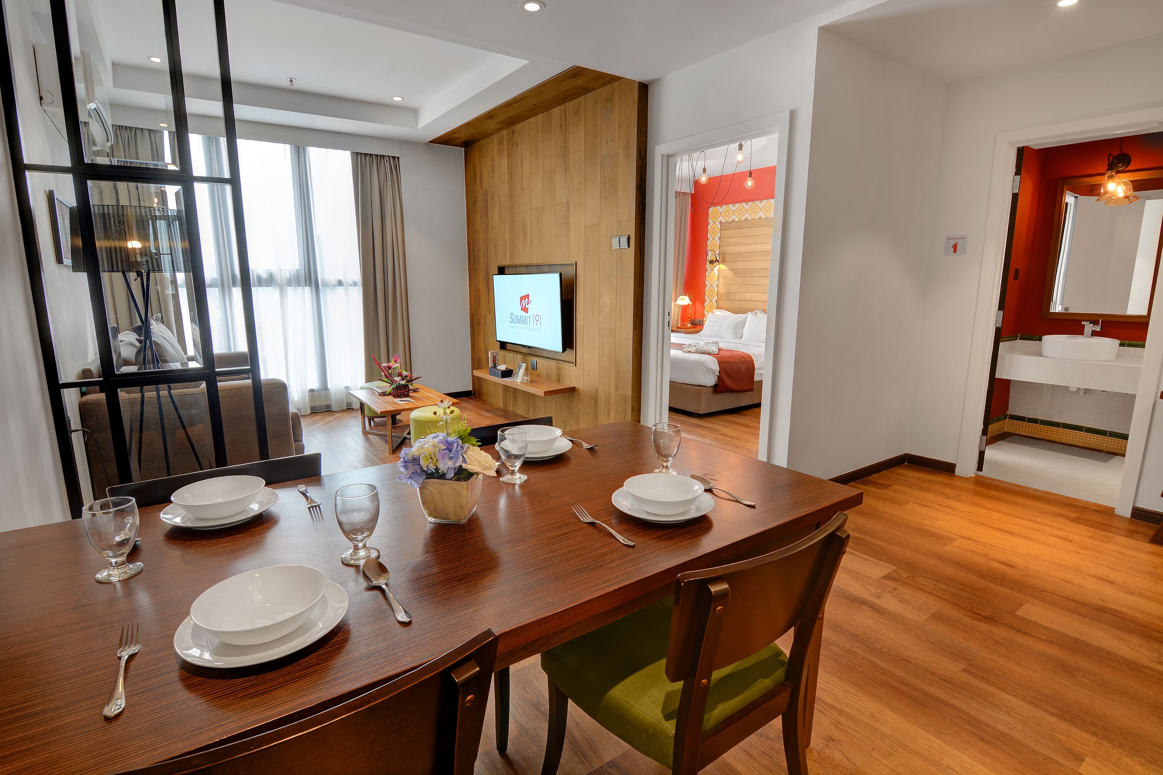 Habitación de hotel con comedor, un televisor mostrando 'Summit 19', cama y baño con espejo y lavamanos.