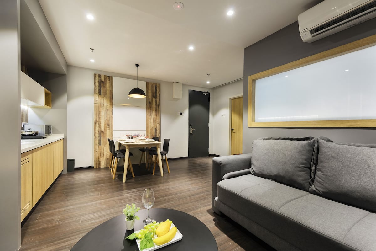 A modern living room with a grey sofa, a dining area with a wooden table and chairs, and a kitchen area with wooden cabinets.