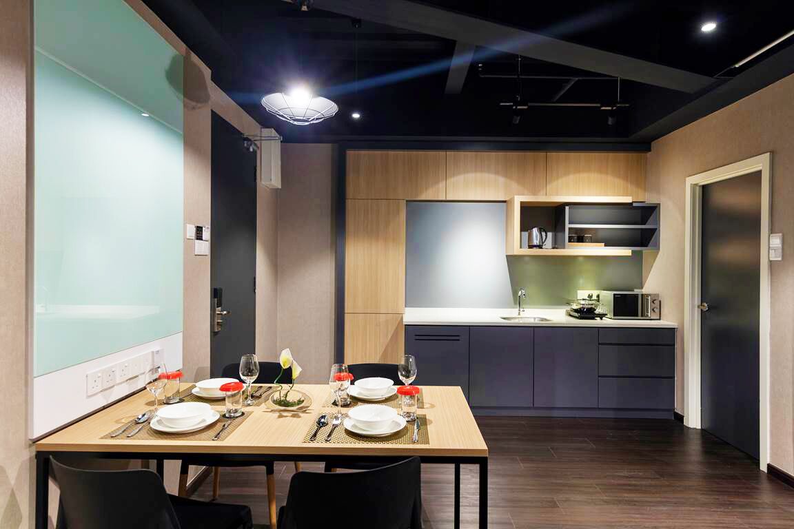 A modern dining area with a wooden table set for two, a kitchen in the background, and a light fixture above.