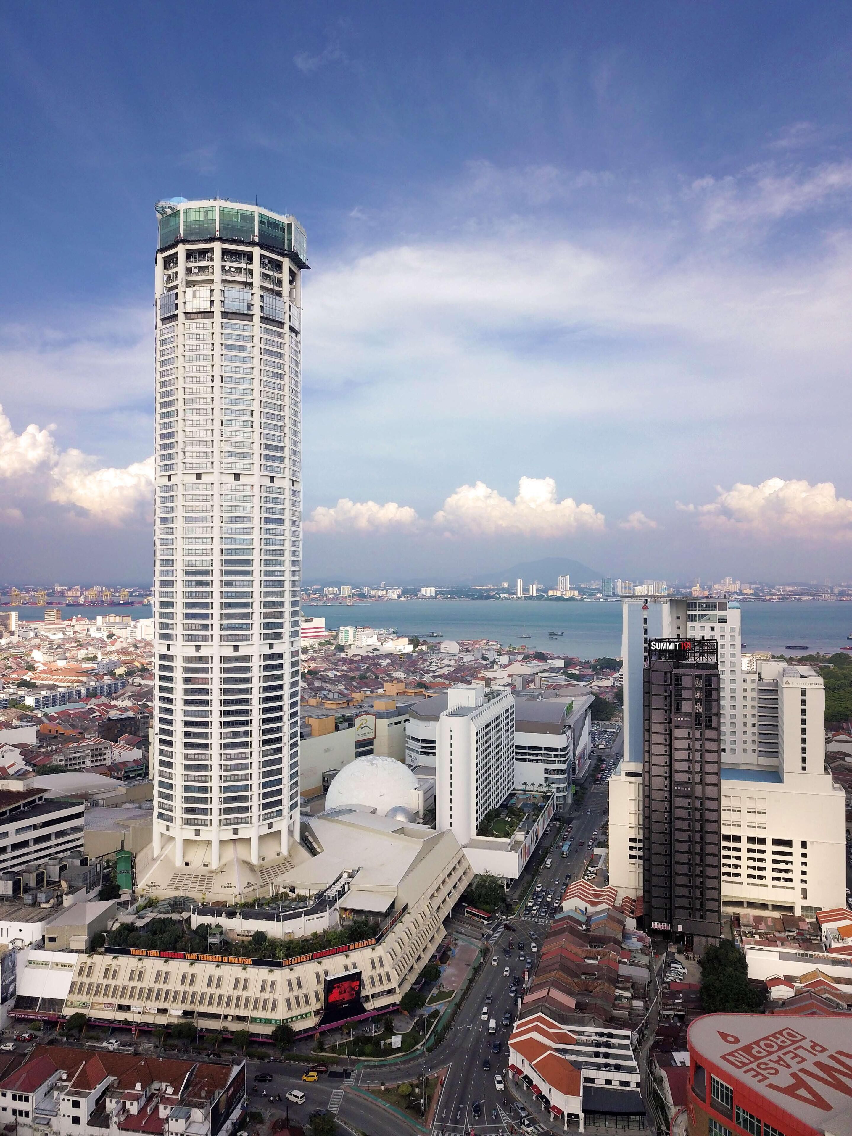 A tall, cylindrical skyscraper with a glass facade, surrounded by a cityscape with various buildings and a body of water in the background.