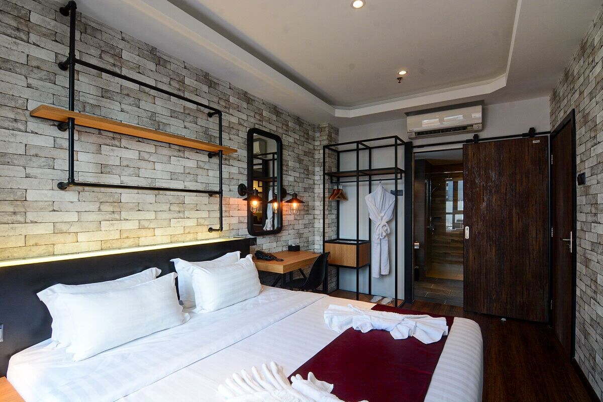 A modern hotel room with a brick wall, a bed with white linens, a wooden desk, and a robe rack.