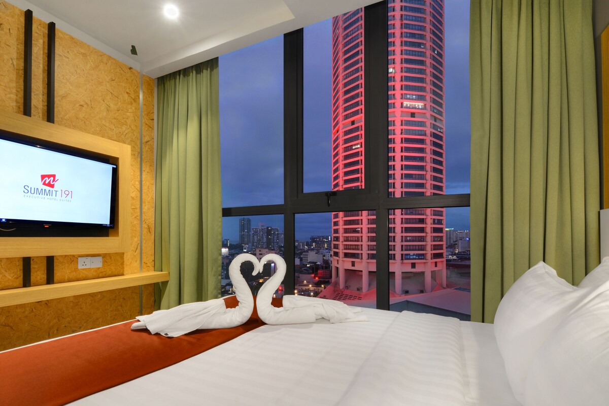 A hotel room with a large window, a bed, a TV displaying 'Summit 191', and a heart-shaped towel arrangement.