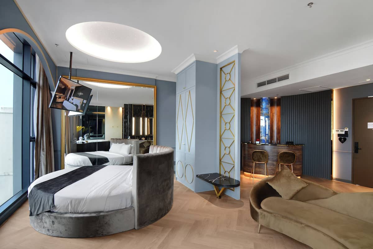 A modern hotel room with a curved bed, a sofa, a dining area with a bar, and a large mirror.