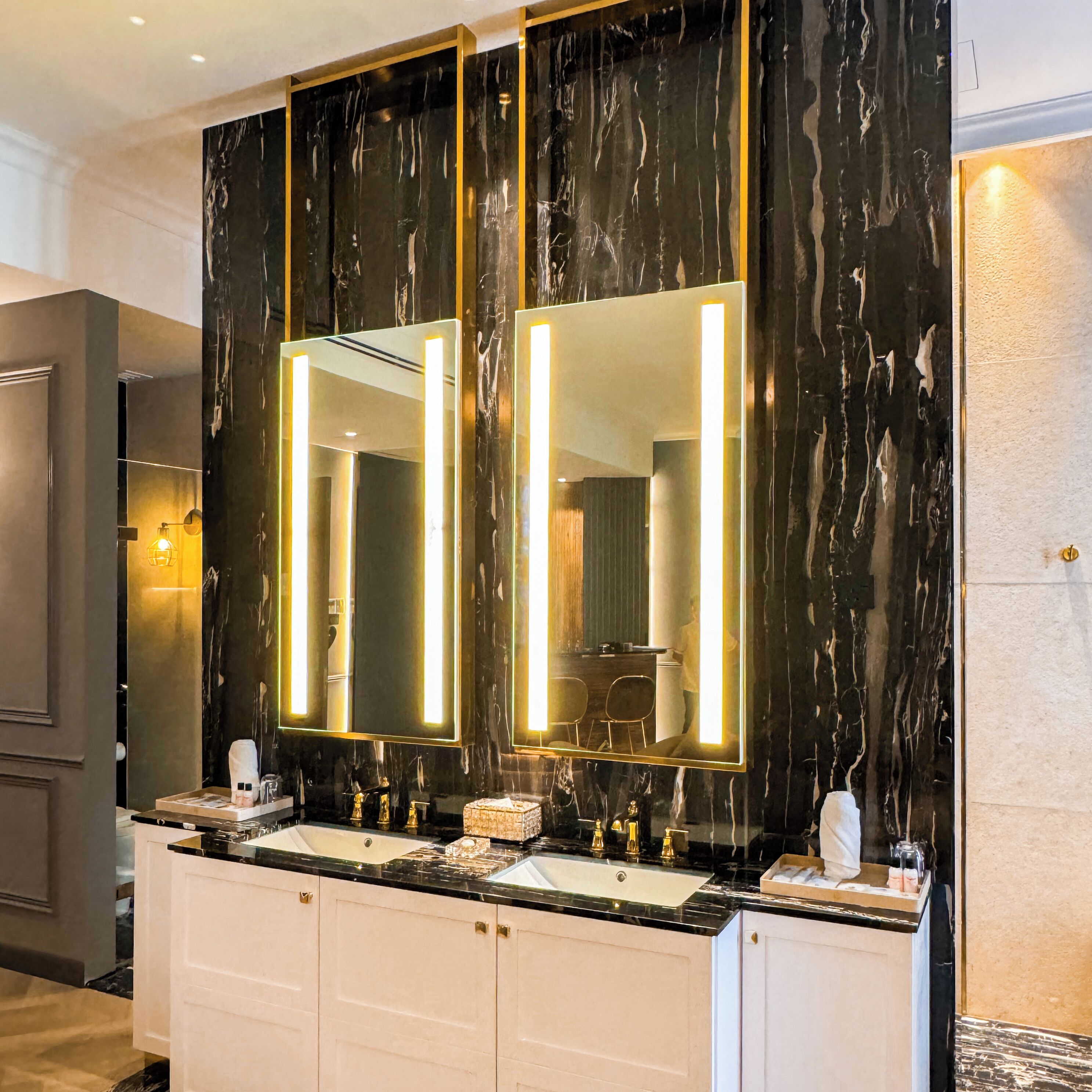 A modern bathroom with a dark marble backsplash, two large mirrors with illuminated frames, a white vanity with a black countertop, and a sink with gold fixtures.