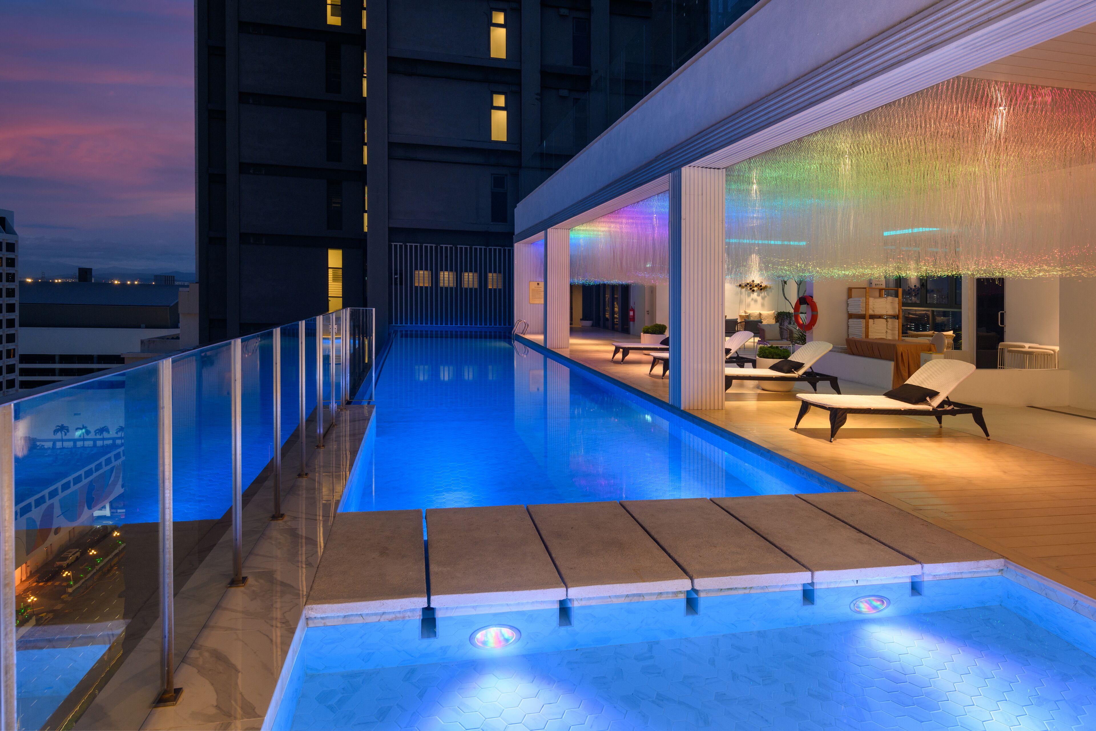 A rooftop pool area with a glass balustrade, lounge chairs, and a view of the cityscape at dusk.