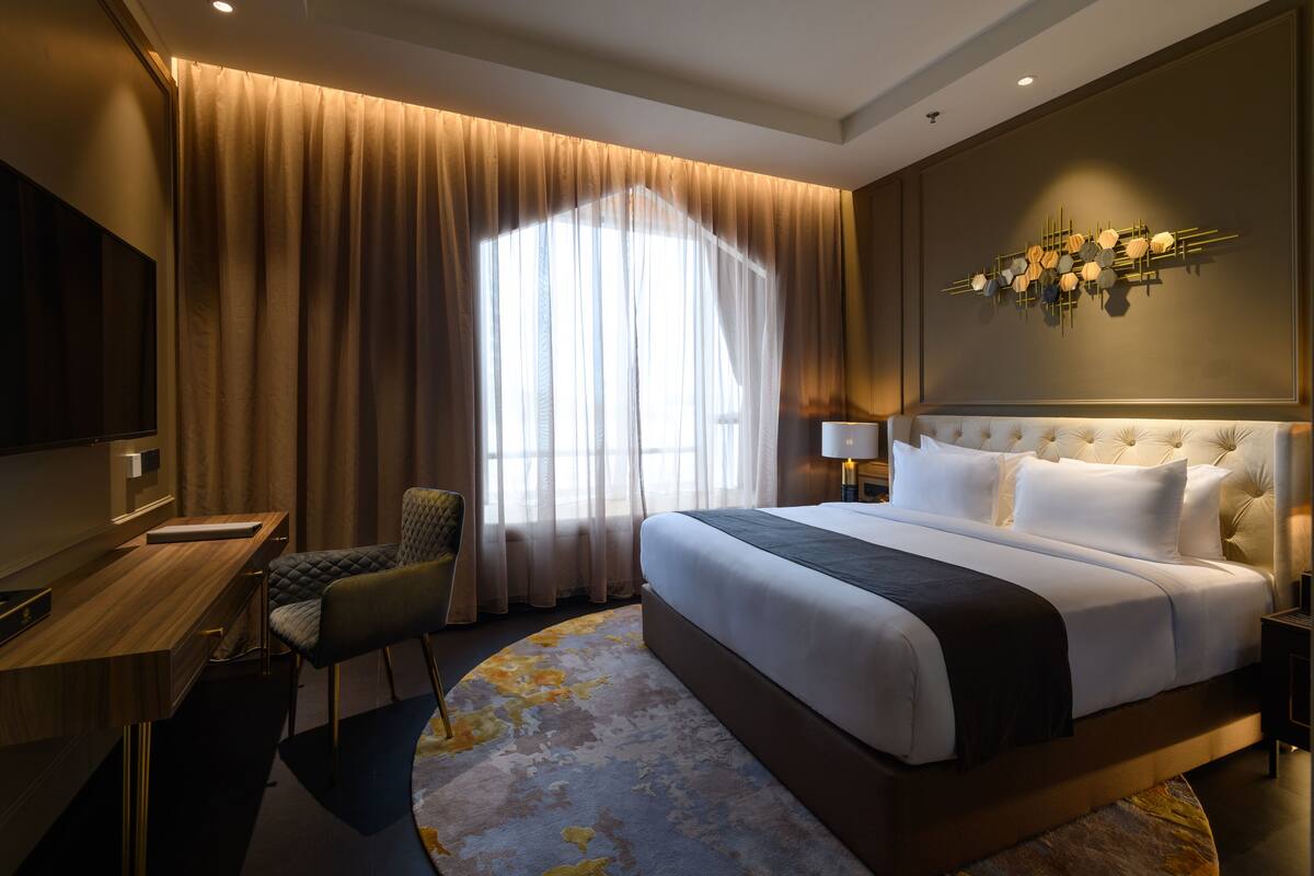 A hotel room with a large bed, a desk, a chair, a TV, and a window with curtains.