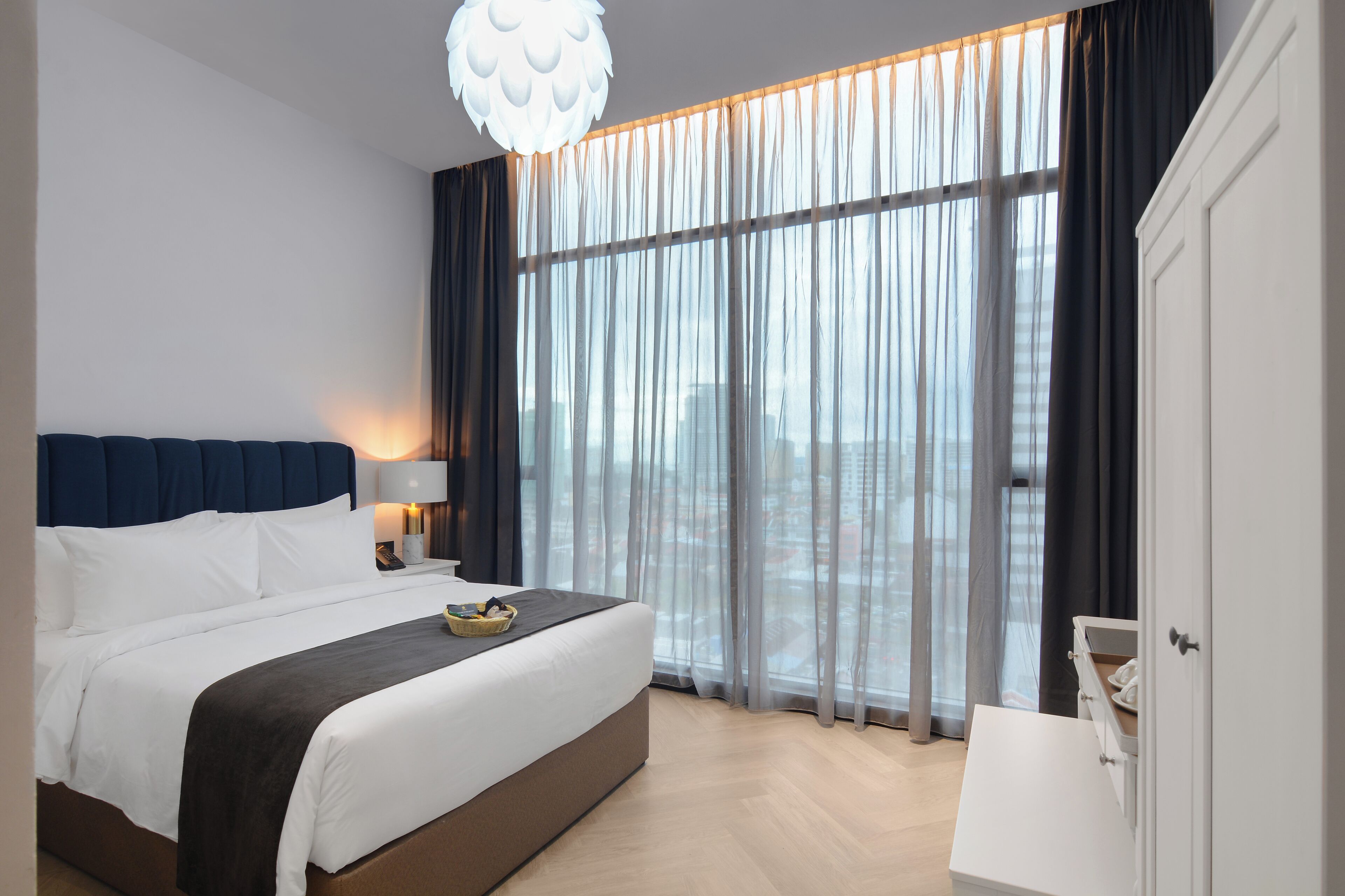 A modern hotel room with a large bed, a bedside table with a lamp, a view of the city through sheer curtains, and a white wardrobe.