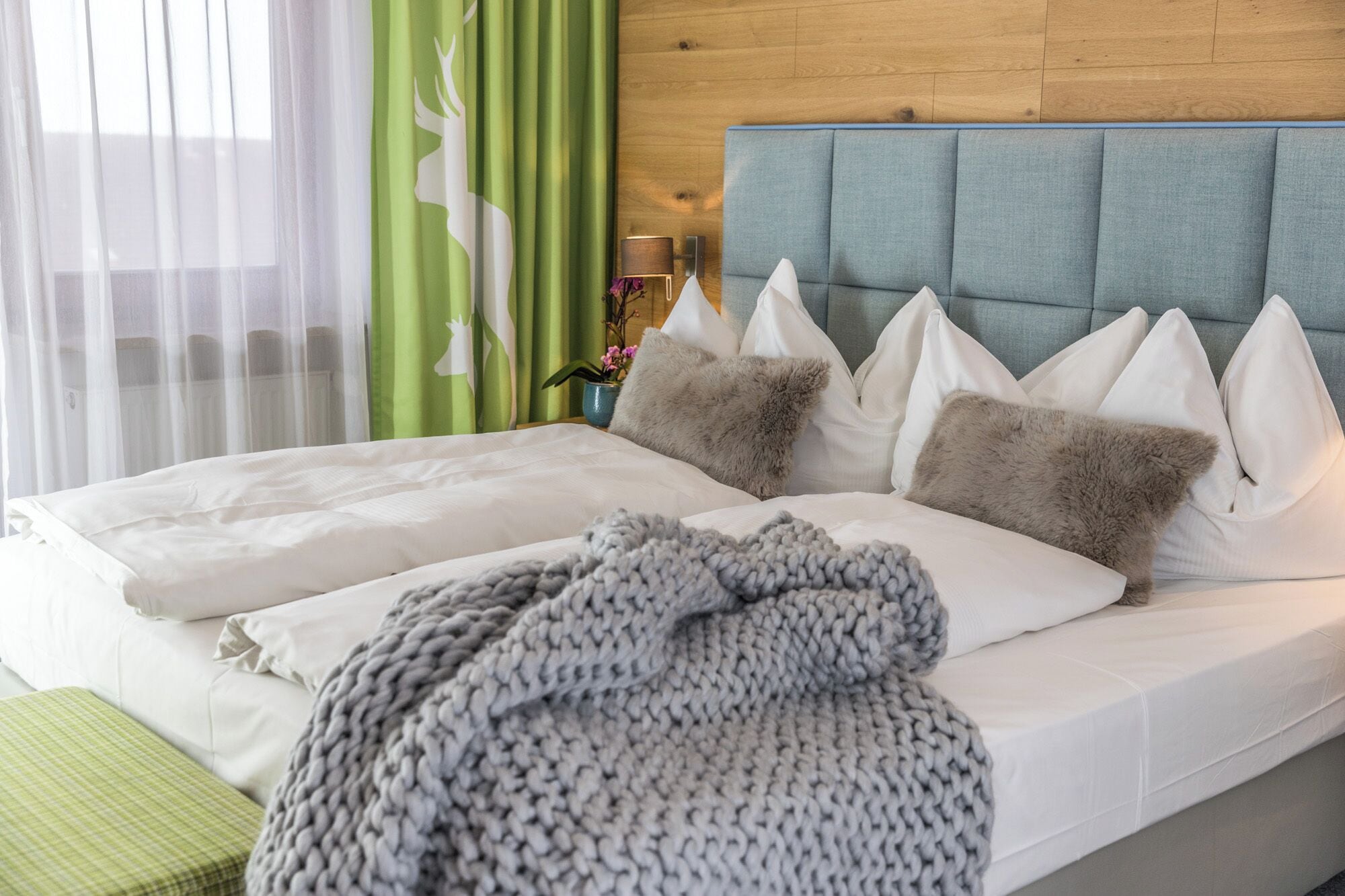 Una cama con sábanas blancas, un plaid de punto gris y varias almohadas.
