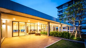 A modern building with a covered outdoor area featuring wooden flooring, glass doors, and a seating area. - Burgundy Residence (Si Maha Phot)