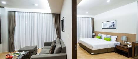 Una habitación de hotel moderna con un sofá, una mesita con revistas, una cama con almohadas, una mesita de noche con lámpara y un ventanal con cortinas.