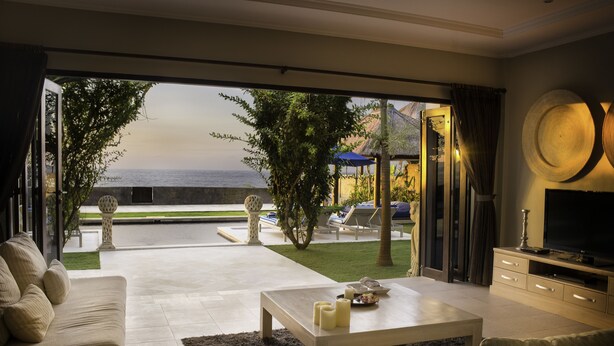 A modern living room with a large glass door leading to a pool area, a flat-screen TV, and a dining table with candles. - The Beach Front Villas - North Bali (Sawan)