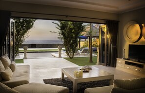 A modern living room with a large glass door leading to a pool area, a flat-screen TV, and a dining table with candles. - The Beach Front Villas - North Bali (Sawan)