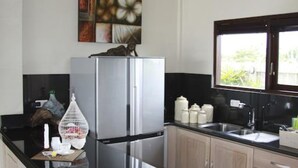A modern kitchen with a black countertop, stainless steel refrigerator, wooden cabinets, and a ceiling fan. - The Beach Front Villas - North Bali (Sawan)