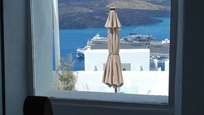 A view from a window overlooking a coastal area with a chair and an umbrella outside. - Akron Cliff Suite (Santorini)