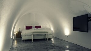 A bedroom with a bed, bedside table, and a flat-screen TV mounted on the wall. - Akron Cliff Suite (Santorini)