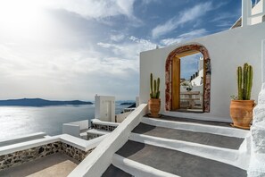 A white building with a stone base, a wooden door, and a view of the sea. - Akron Cliff Suite (Santorini)