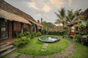 Une maison traditionnelle à toit de chaume, dotée d’une petite piscine circulaire entourée d’un chemin de pierre et d’une végétation luxuriante.