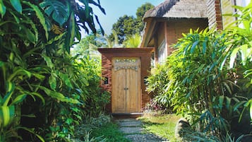 Caminho que leva a uma porta de madeira com uma placa, cercado por vegetação exuberante e um edifício de tijolos.