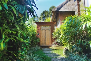 Un sentier menant à une porte en bois, sur laquelle est apposée une plaque, le tout entouré d’une végétation luxuriante et d’un bâtiment en briques.