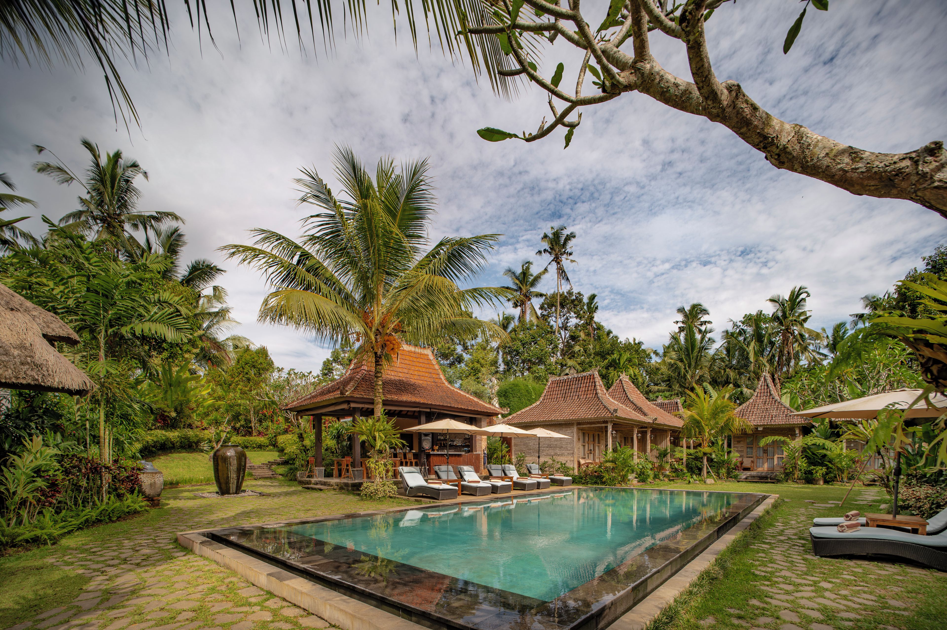 A pool area with lounge chairs, a thatched-roof structure, and tropical vegetation.
