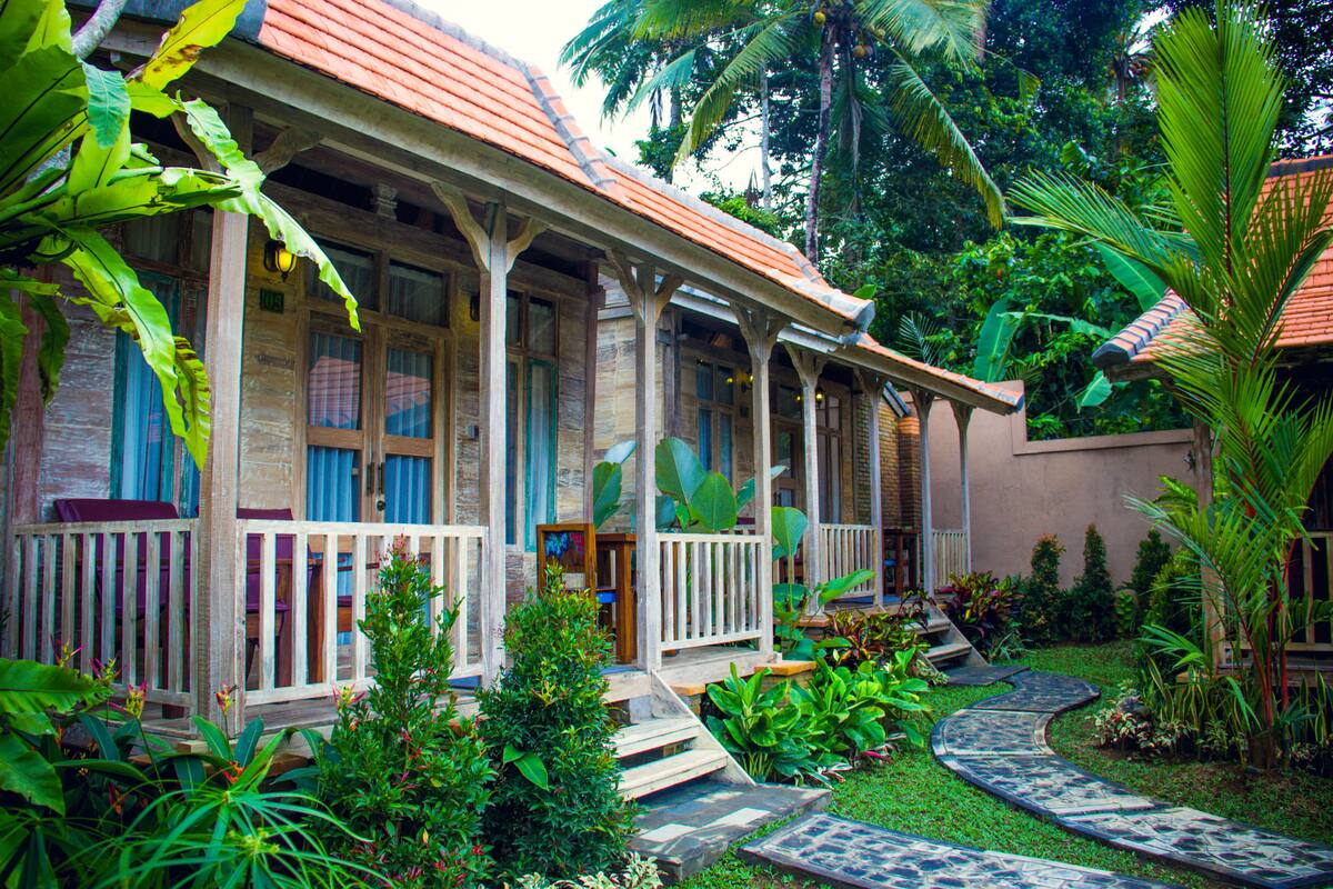 Une maison traditionnelle en bois, avec un toit de tuiles, entourée d’une végétation luxuriante et bordée d’un chemin pavé de pierres.