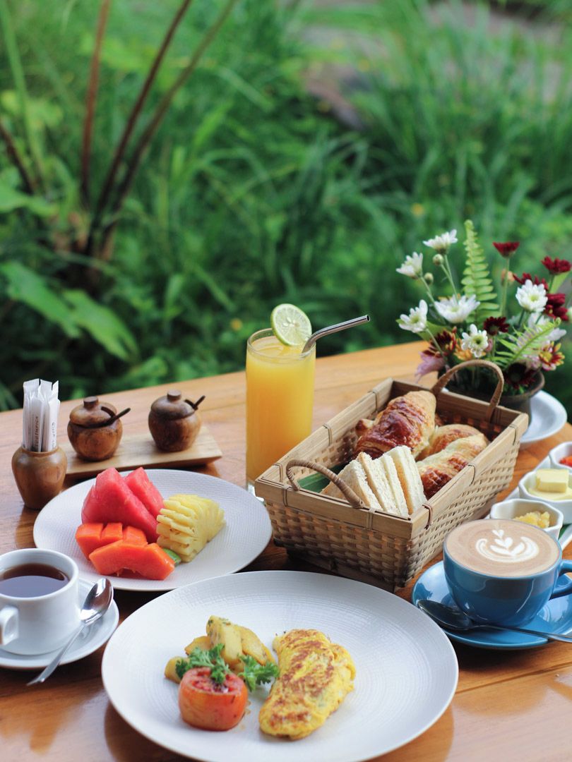 A breakfast setting with a variety of food items, including fruits, pastries, and beverages, arranged on a wooden table with a natural backdrop.