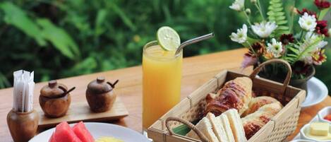 A breakfast setting with a variety of food items, including fruits, pastries, and beverages, arranged on a wooden table with a natural backdrop.