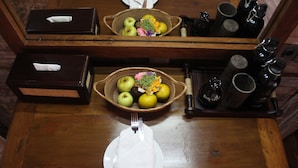 Une table en bois avec une assiette, une serviette pliée, un panier de fruits et une bouteille d’eau.