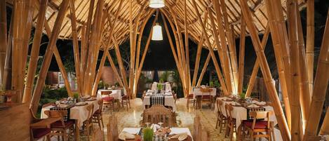 Un comedor interior con estructuras de techo de bambú, mesas dispuestas con manteles blancos y sillas.