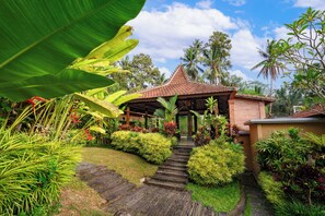 Un bâtiment traditionnel avec un toit en tuiles, entouré d’une végétation tropicale luxuriante et accessible par un chemin de pierre.