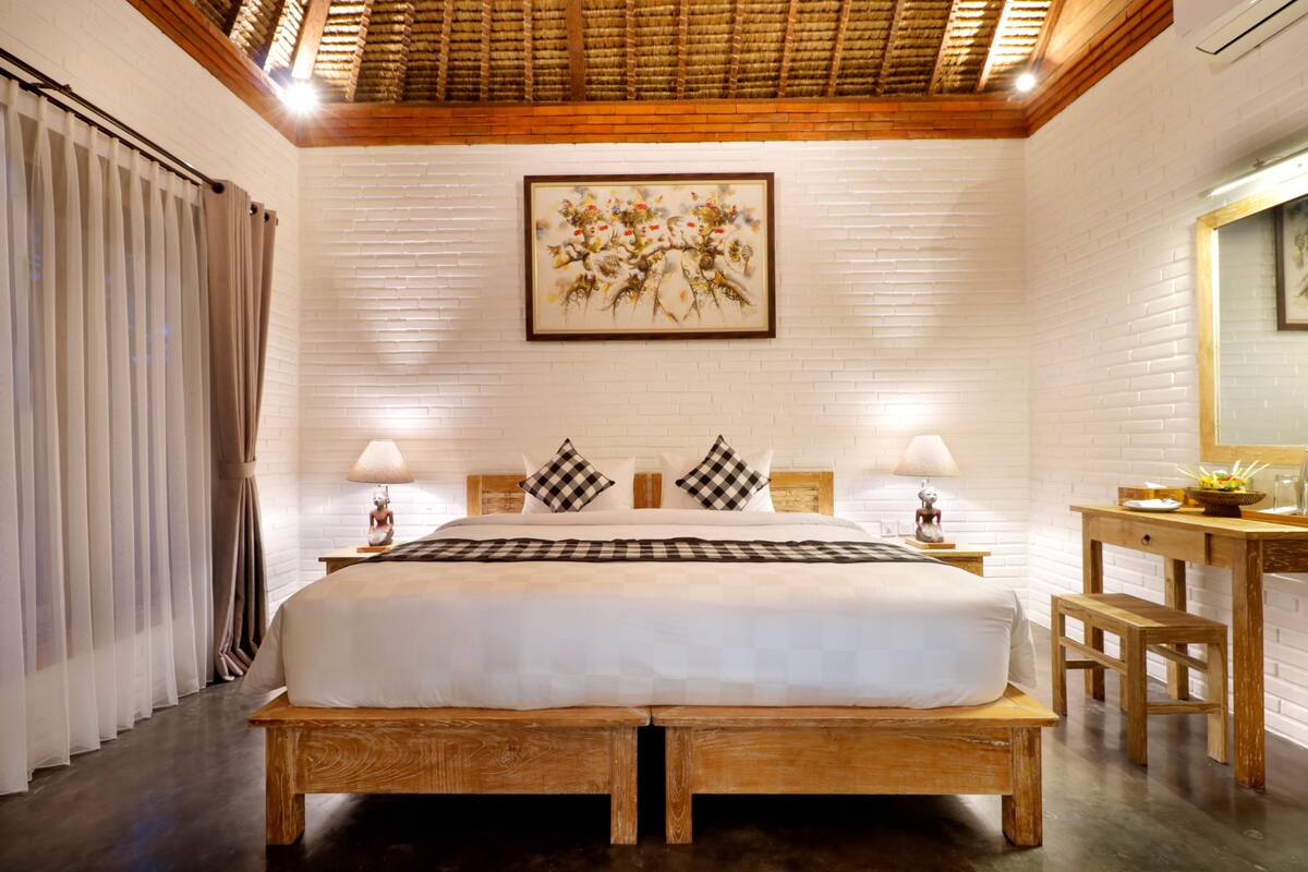 Une chambre spacieuse avec un grand lit, une tête de lit en bois, deux tables de chevet avec des lampes, un banc en bois, un petit bureau avec un miroir et un mur en briques blanches orné d’une œuvre d’art encadrée au-dessus du lit.