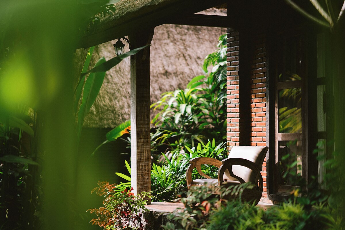 Une chaise en bois placée sous un toit de chaume, entourée d’une végétation luxuriante et d’un mur de briques.