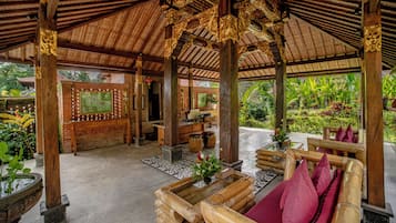 A traditional wooden structure with intricate carvings, a seating area with cushions, and a view of lush greenery outside.