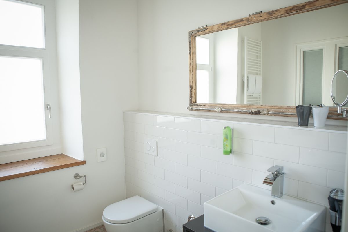 Un baño con inodoro blanco, lavamanos con espejo y ventana con estante de madera.
