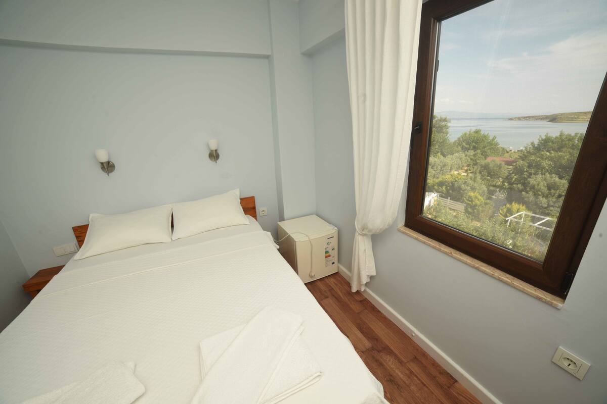 A bedroom with a bed, a nightstand, a window with a view of greenery and water, and a wall-mounted light fixture.