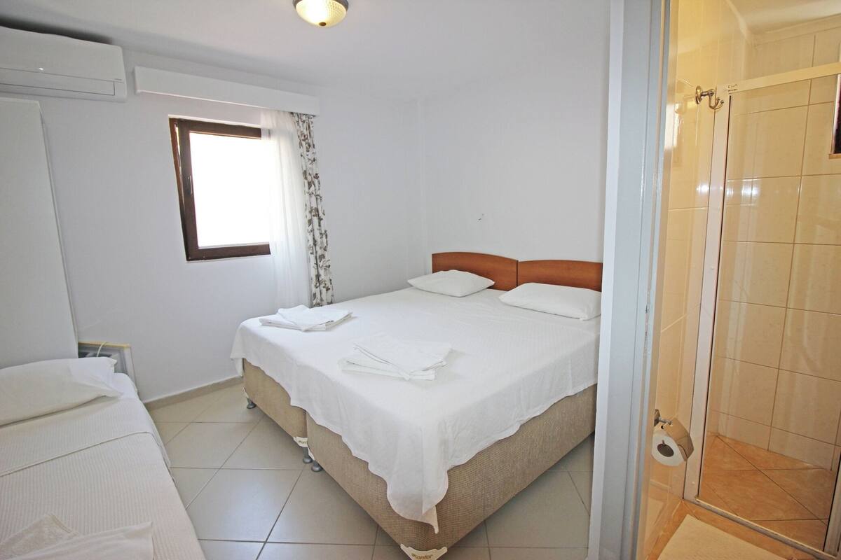 A bedroom with a bed, a window with curtains, a shower area, and a door.