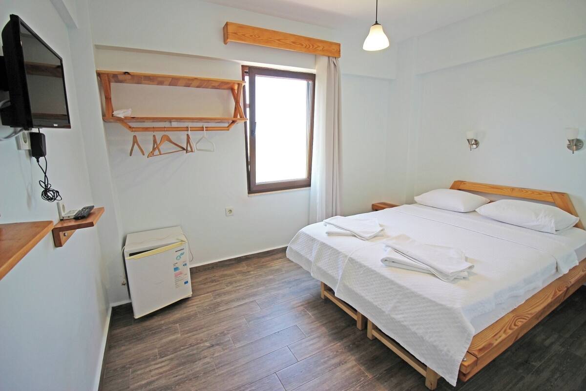 A bedroom with a bed, a TV mounted on the wall, a heater, a window with curtains, and a wooden shelf with hangers.