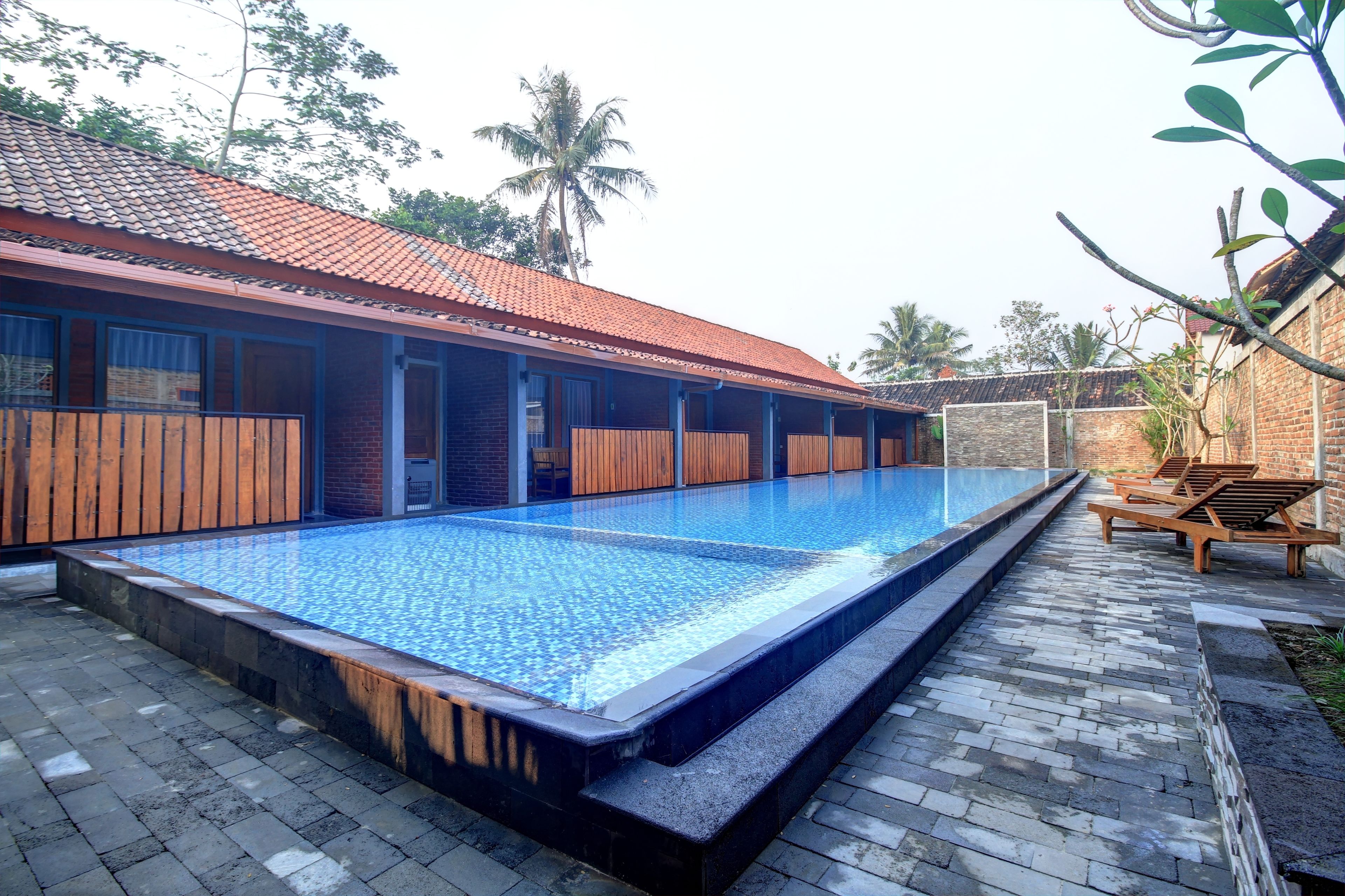 Uma piscina com um deck ladrilhado, cadeiras de madeira e um edifício com telhado telhado.