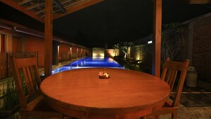 A wooden table with chairs in a covered outdoor area with a pool in the background.
