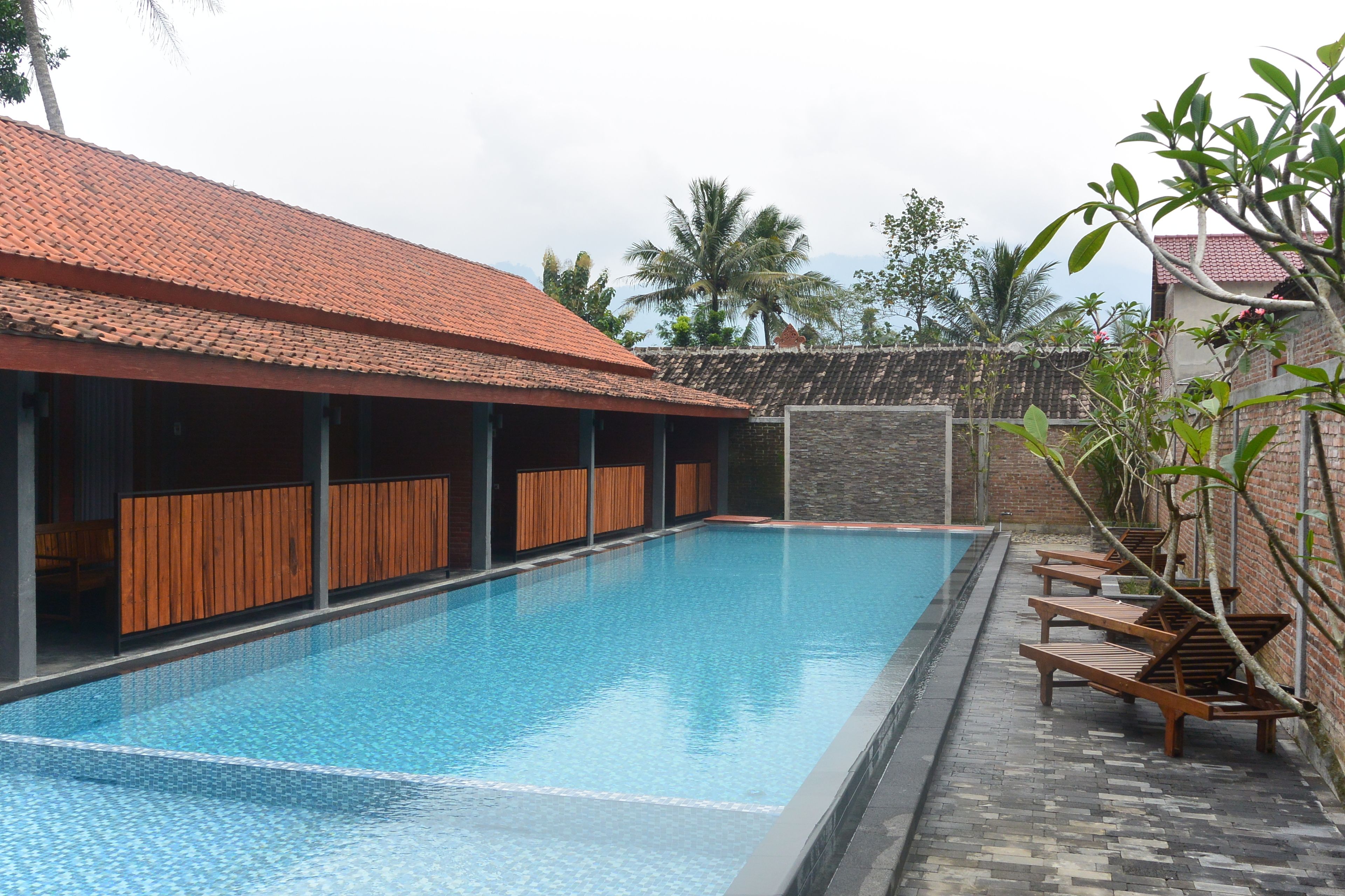A swimming pool with wooden deck chairs, a tiled roof, and a stone wall.