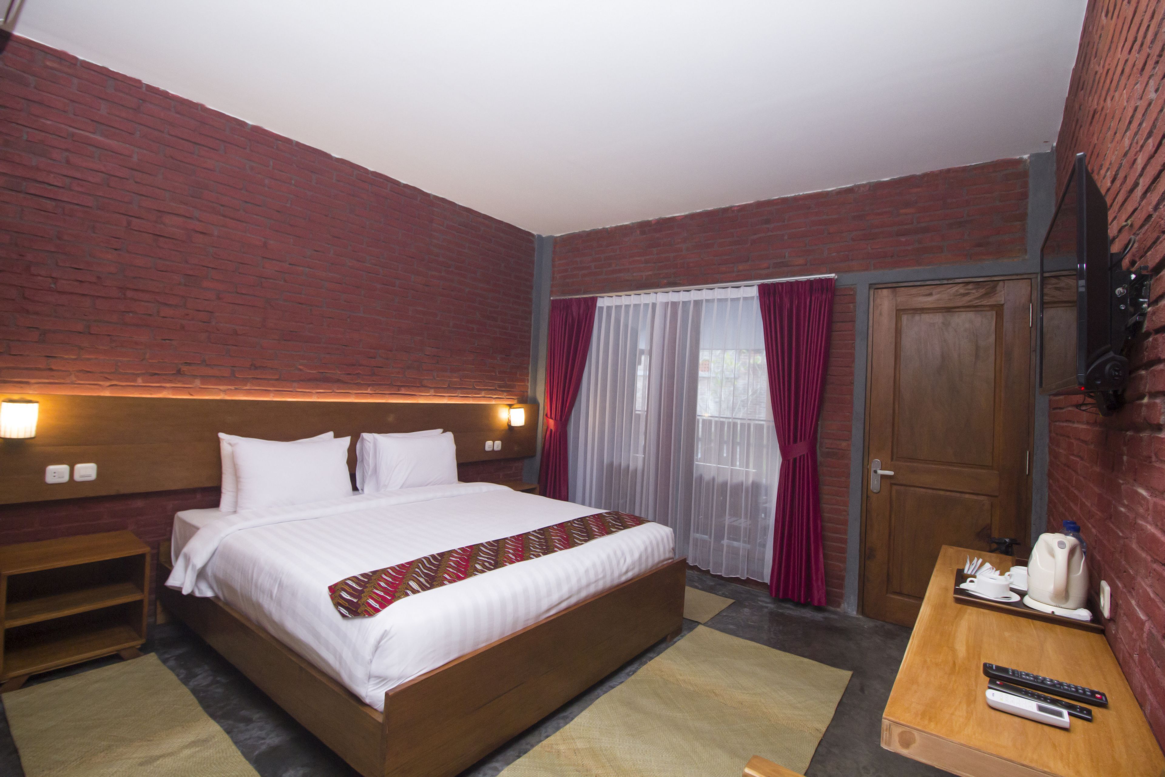 A hotel room with a large bed, a wooden nightstand, a desk with a kettle and remote controls, a brick wall, and a window with curtains.