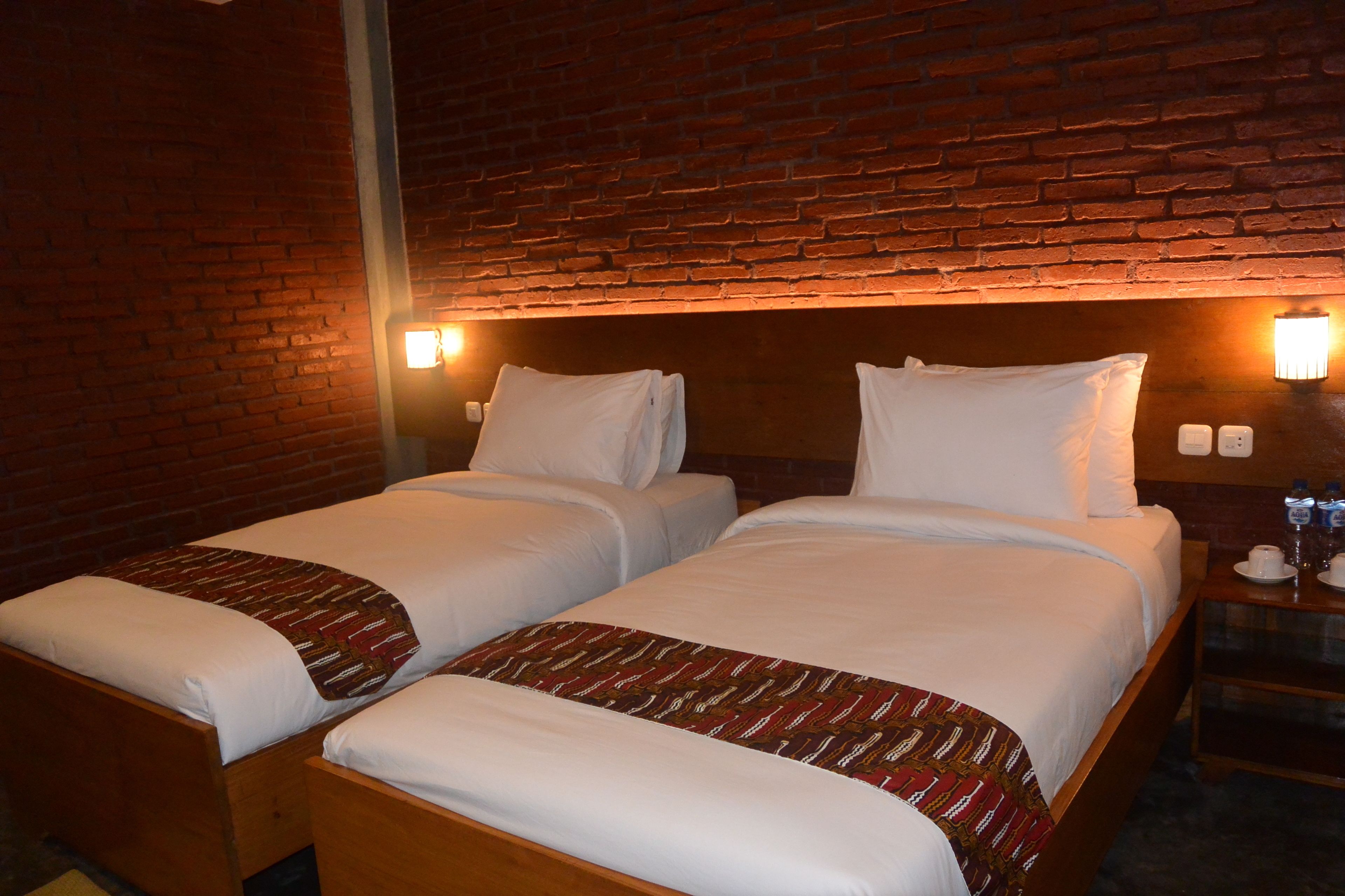 A hotel room with two beds, a brick wall, and a nightstand with a lamp and a cup.