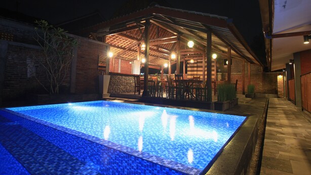 A swimming pool with a tiled edge, a covered patio area with chairs, and a brick wall.