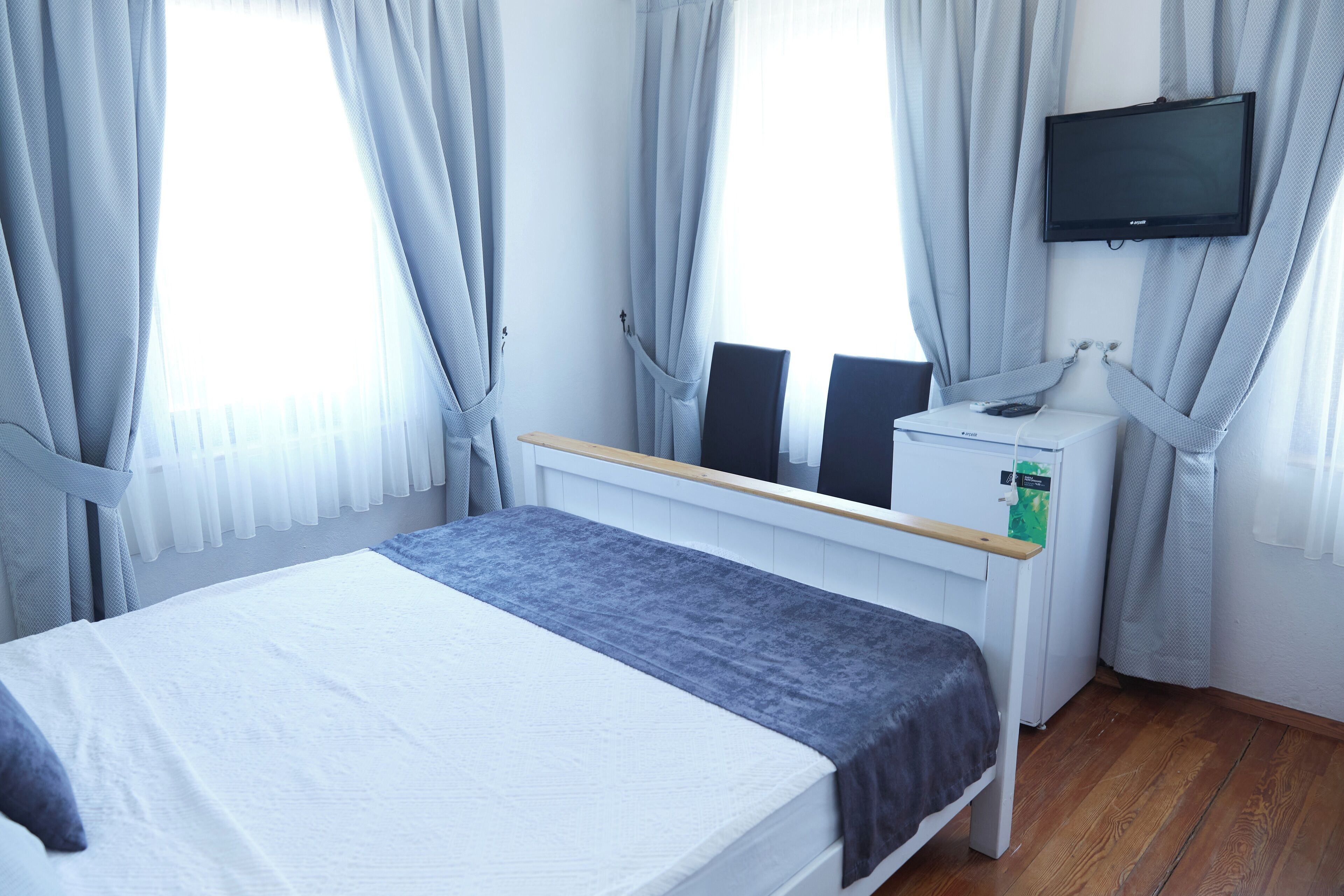 A bedroom with a bed, a desk with a computer, a TV mounted on the wall, and a window with curtains.