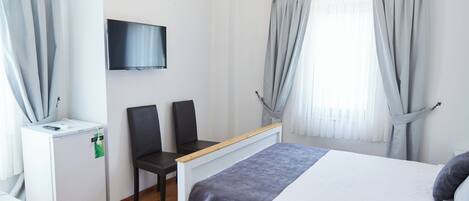 Una habitación de hotel con cama, escritorio, silla, televisor y ventana con cortinas.