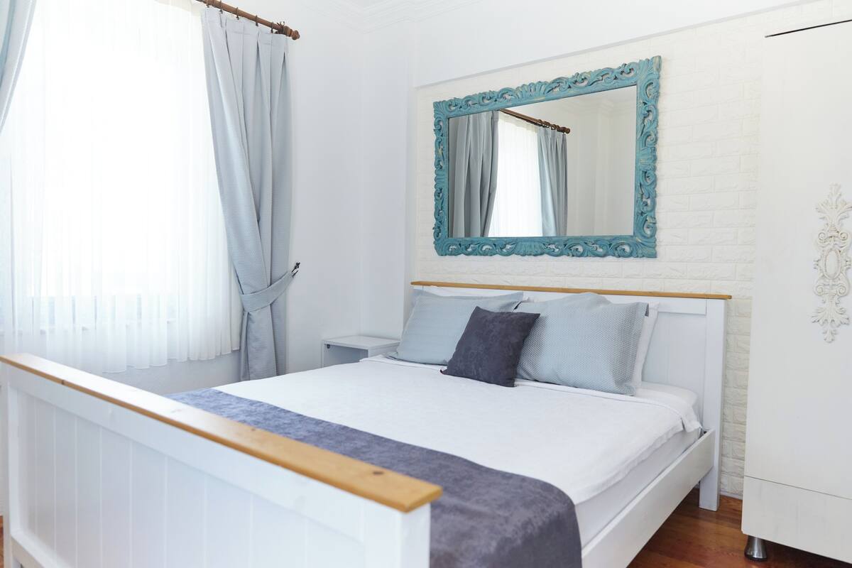 A bedroom with a white bed, a wooden headboard, a nightstand, a mirror with a blue frame, and a white wall with a decorative molding.