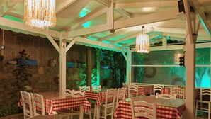 An indoor dining area with tables covered in red and white checkered tablecloths, white chairs, and hanging chandeliers. - Ebruli Hotel (Bozcaada)