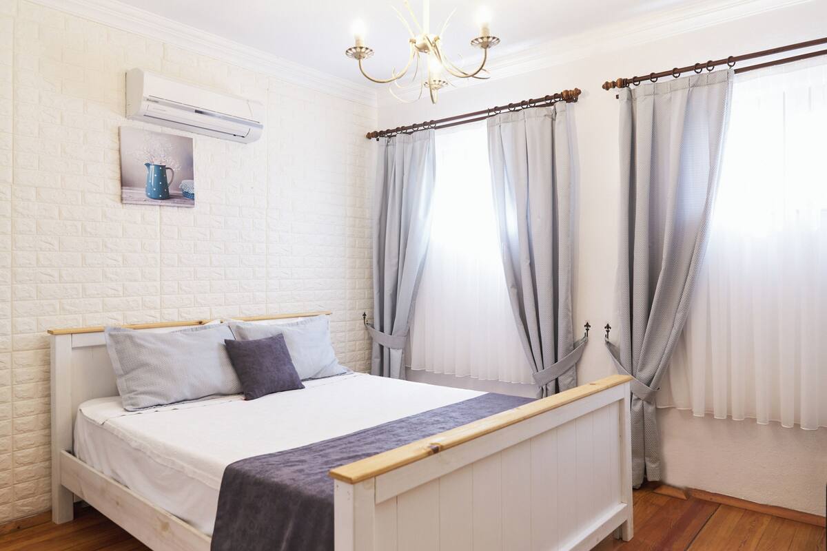 A bedroom with a white brick wall, a bed with a wooden headboard, a white air conditioner, and a chandelier.